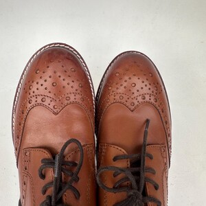 May include: A pair of brown leather Oxford shoes with decorative perforations and dark brown laces. The shoes have a classic wingtip design and a stitched sole. The leather has a polished finish, and the shoes are set against a white background.