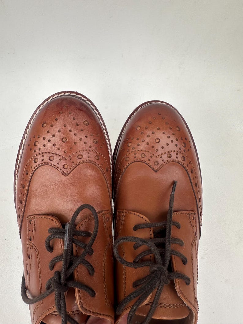 May include: A pair of brown leather Oxford shoes with decorative perforations and dark brown laces. The shoes have a classic wingtip design and a stitched sole. The leather has a polished finish, and the shoes are set against a white background.