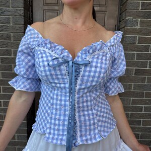 Puede incluir: Un top estilo cors&eacute; de cuadros vichy en azul claro y blanco con un peque&ntilde;o estampado floral, mangas abullonadas ca&iacute;das con ribete de encaje blanco y un cierre central de cordones con cinta azul. La prenda se combina con una falda blanca.