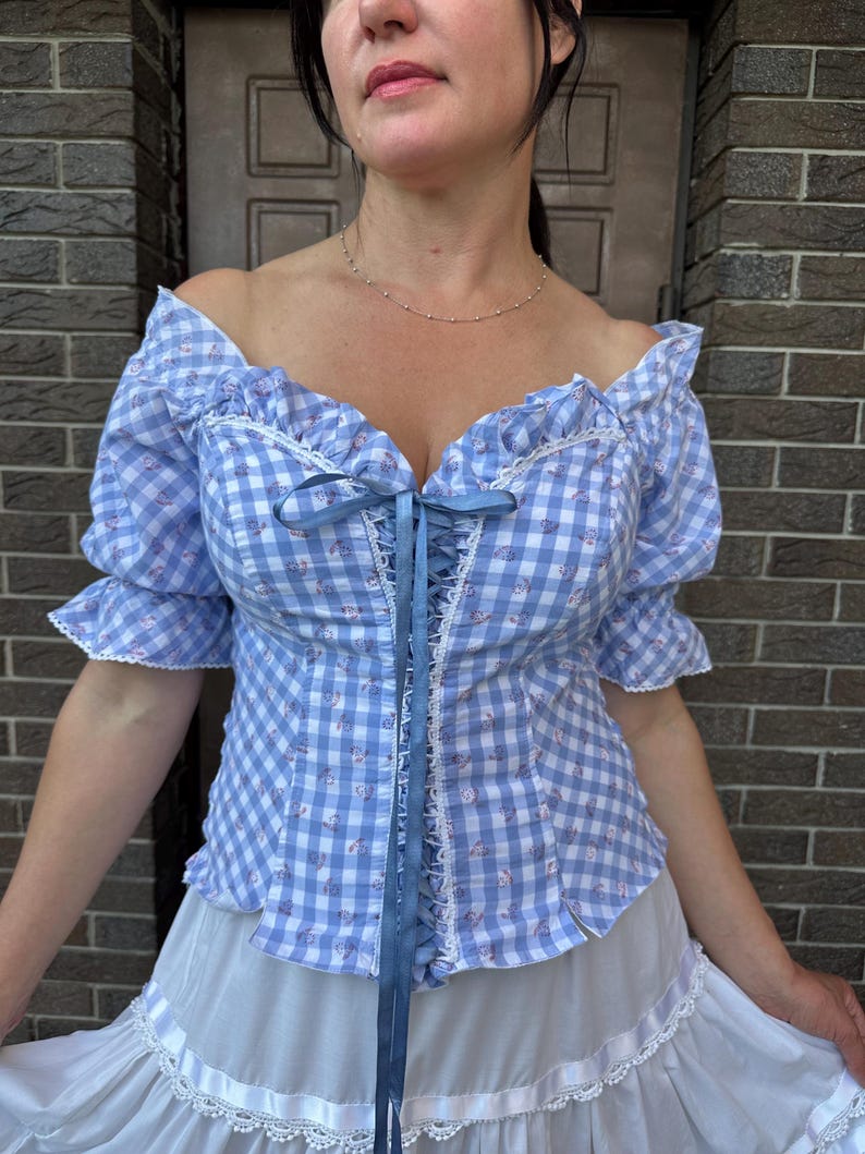 Puede incluir: Un top estilo cors&eacute; de cuadros vichy en azul claro y blanco con un peque&ntilde;o estampado floral, mangas abullonadas ca&iacute;das con ribete de encaje blanco y un cierre central de cordones con cinta azul. La prenda se combina con una falda blanca.