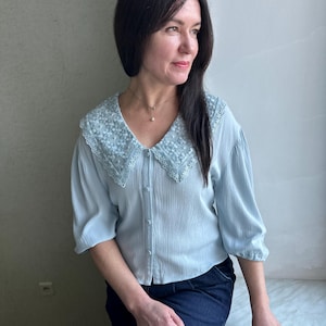 Puede incluir: Una blusa azul claro con un cuello detallado con encaje. La blusa tiene mangas tres cuartos y una botonadura. La modelo lleva vaqueros azul oscuro. La blusa es de un estilo suave y femenino.