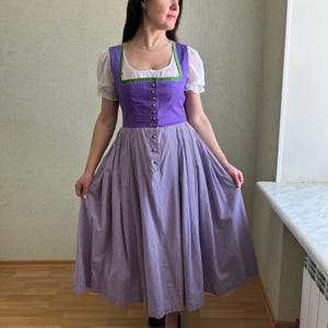 May include: A traditional purple dirndl dress with a white blouse and a checkered skirt. The bodice is a vibrant purple with green trim and buttons down the front. The skirt is a checkered pattern in purple and white.