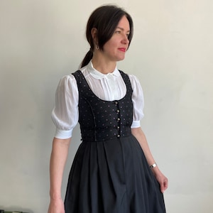 Puede incluir: Un vestido dirndl negro que presenta un corpi&ntilde;o con un sutil patr&oacute;n de puntos morados y una hilera de botones met&aacute;licos, combinado con una blusa blanca de mangas cortas abullonadas, cuello redondo y pliegues verticales en la tapeta frontal.