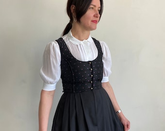 Puede incluir: Un vestido dirndl negro que presenta un corpiño con un sutil patrón de puntos morados y una hilera de botones metálicos, combinado con una blusa blanca de mangas cortas abullonadas, cuello redondo y pliegues verticales en la tapeta frontal.