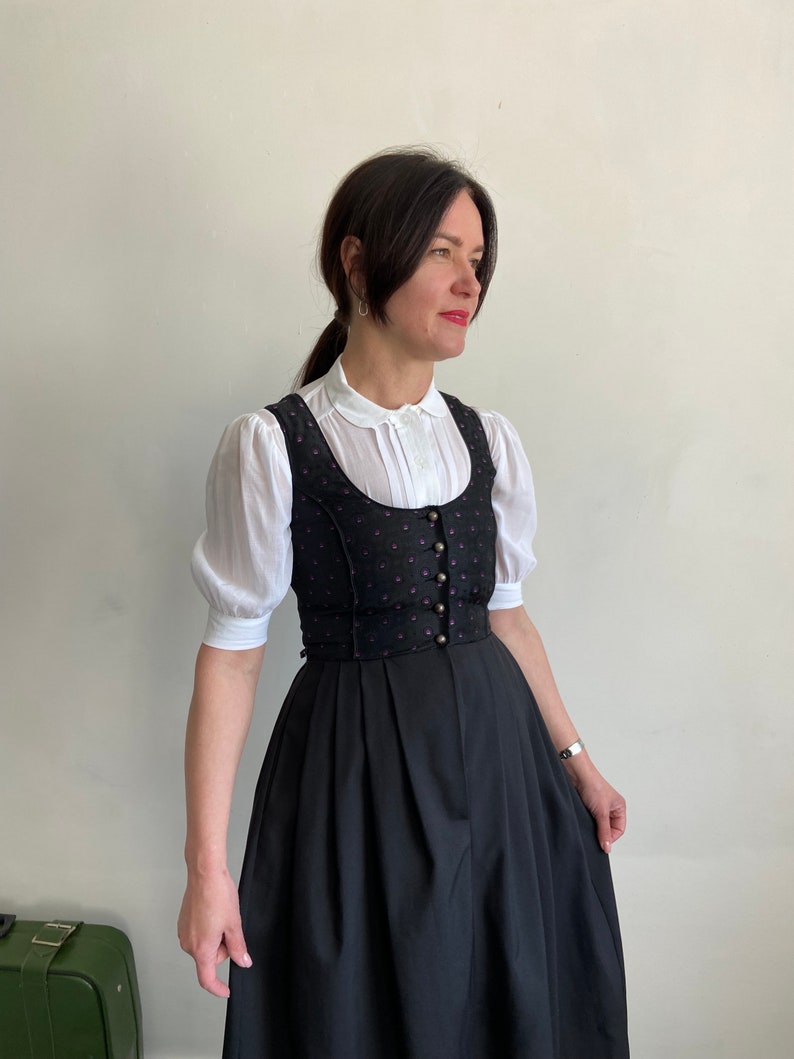 Puede incluir: Un vestido dirndl negro que presenta un corpi&ntilde;o con un sutil patr&oacute;n de puntos morados y una hilera de botones met&aacute;licos, combinado con una blusa blanca de mangas cortas abullonadas, cuello redondo y pliegues verticales en la tapeta frontal.