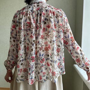 May include: A white long-sleeved blouse with a floral print in shades of red, blue, and green. The blouse has a ruffled collar and a gathered back.
