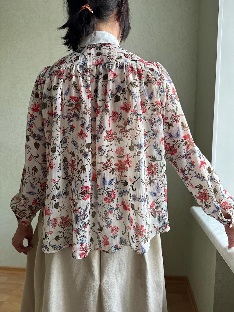 May include: A white long-sleeved blouse with a floral print in shades of red, blue, and green. The blouse has a ruffled collar and a gathered back.