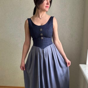 Op de afbeelding: Een blauwe en grijze jurk met een getailleerd bovenstuk en een volle rok. Het bovenstuk heeft een ronde halslijn en is versierd met drie zilveren stervormige knopen. De rok is geplooid bij de taille en valt tot op de grond.