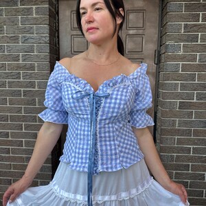 Puede incluir: Una blusa de hombros descubiertos en cuadros vichy azul claro y blanco con mangas cortas con volantes, frente acordonado con cinta azul y detalles florales, combinada con una falda larga blanca de niveles con encaje y cintas.