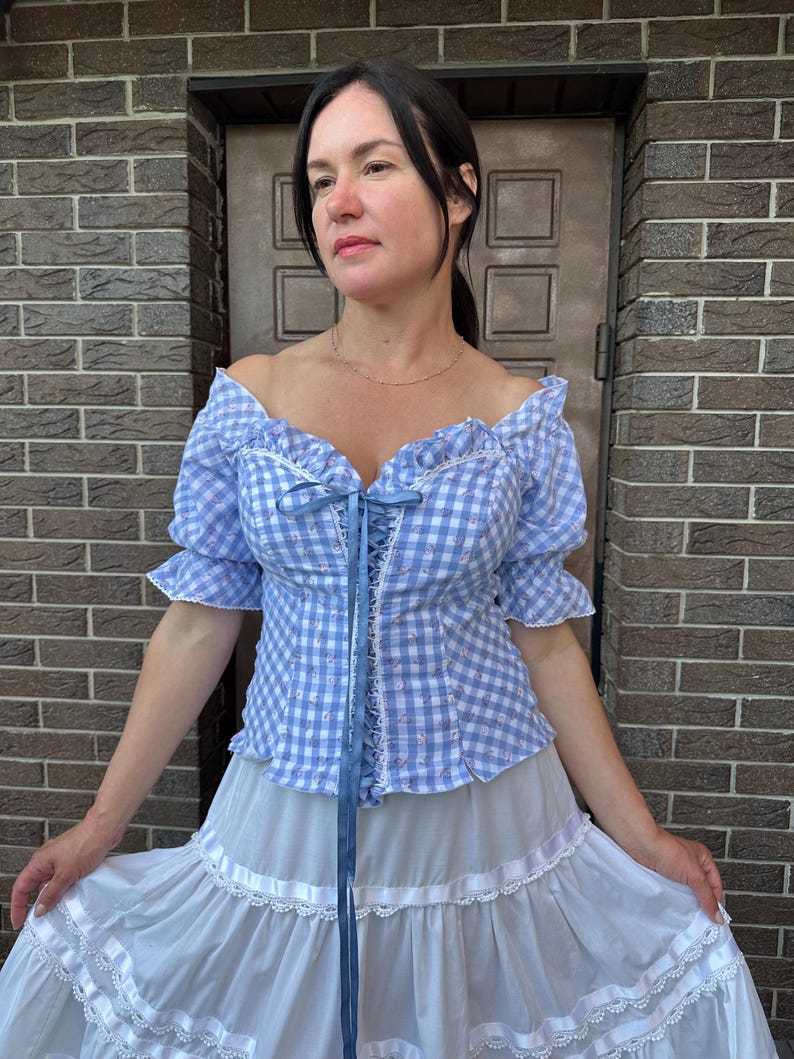 Puede incluir: Una blusa de hombros descubiertos en cuadros vichy azul claro y blanco con mangas cortas con volantes, frente acordonado con cinta azul y detalles florales, combinada con una falda larga blanca de niveles con encaje y cintas.