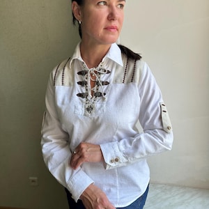 May include: White long-sleeve shirt with decorative brown stitching and toggle closures at the neckline. The shirt features a collared neckline and buttoned cuffs. The shoulders have a beige fabric detail.