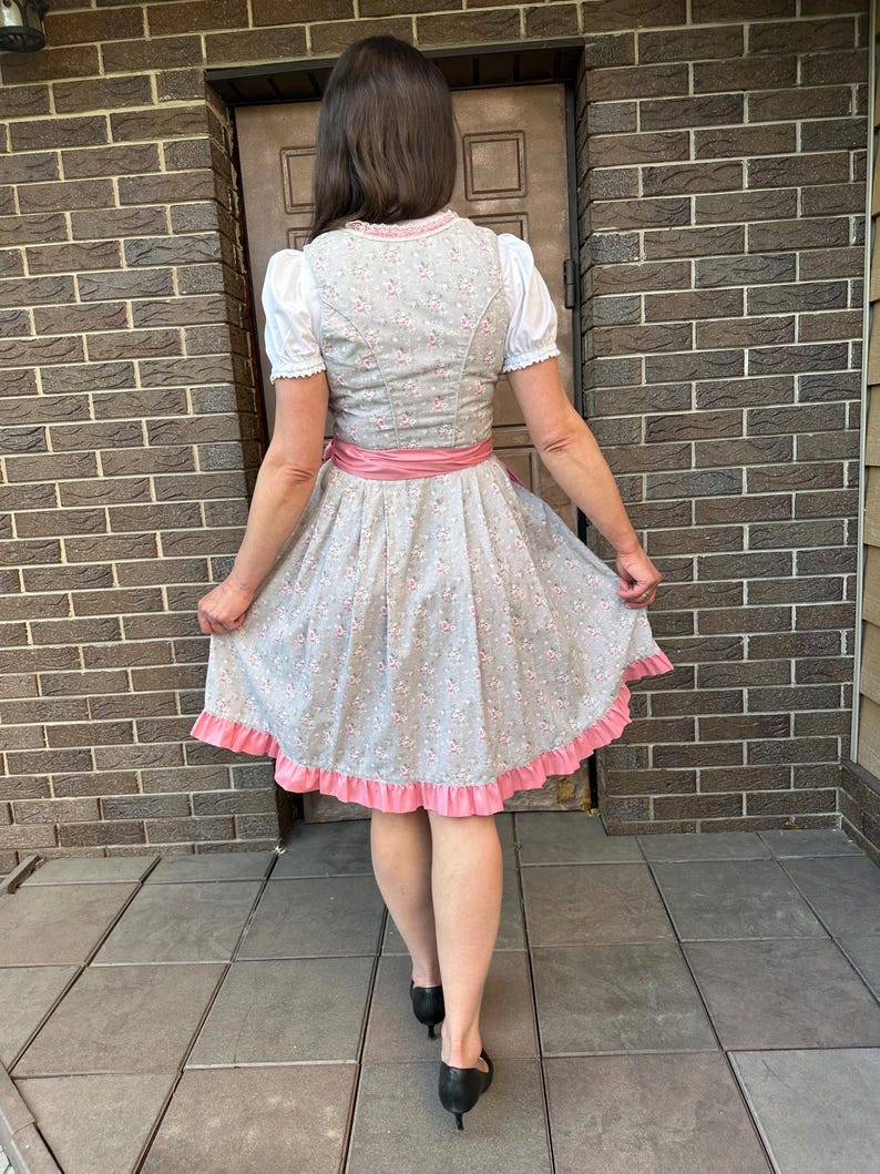 May include: A grey dirndl dress featuring a small pink floral pattern, a white blouse with short puffed sleeves, a solid pink satin waist sash, and a wide pink ruffled hem. The bodice has a high back and the skirt is pleated at the waistline.