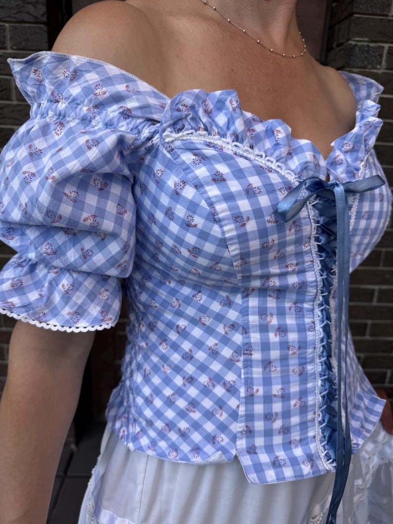Puede incluir: Una blusa de hombros descubiertos con estampado de cuadros vichy en azul claro y blanco, peque&ntilde;os motivos florales, mangas con volantes y cierre frontal de cordones con una cinta azul. Incluye ribetes de encaje blanco en el cuello y mangas.