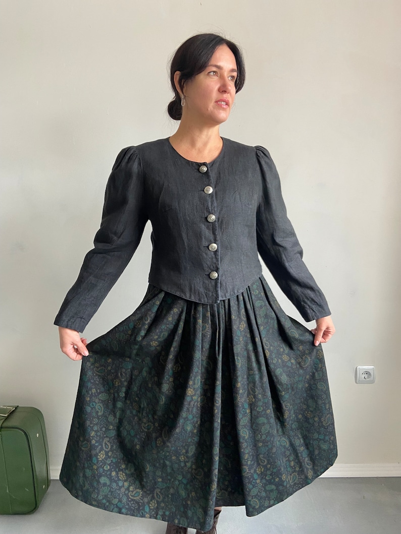 Puede incluir: Una chaqueta negra de lino con mangas abullonadas y botones plateados, puesta sobre una falda negra con estampado paisley verde y azul.