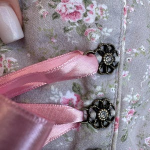 May include: A light gray bodice featuring a floral pattern with pink and white blossoms. The garment is accented with metallic, flower-shaped eyelets in an antique bronze finish, through which a smooth, light pink satin ribbon is laced.
