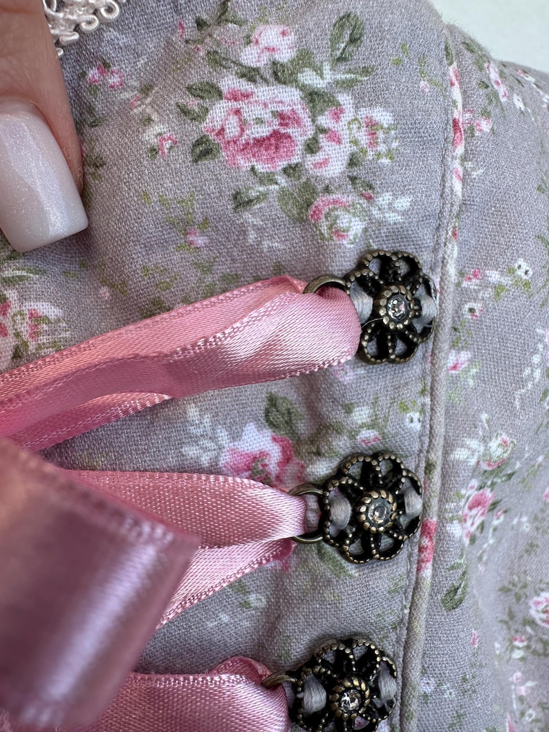 May include: A light gray bodice featuring a floral pattern with pink and white blossoms. The garment is accented with metallic, flower-shaped eyelets in an antique bronze finish, through which a smooth, light pink satin ribbon is laced.