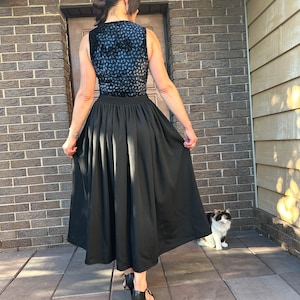 May include: A woman wearing a black skirt with a blue and black patterned top. She is standing on a tiled surface with a cat sitting in the background.