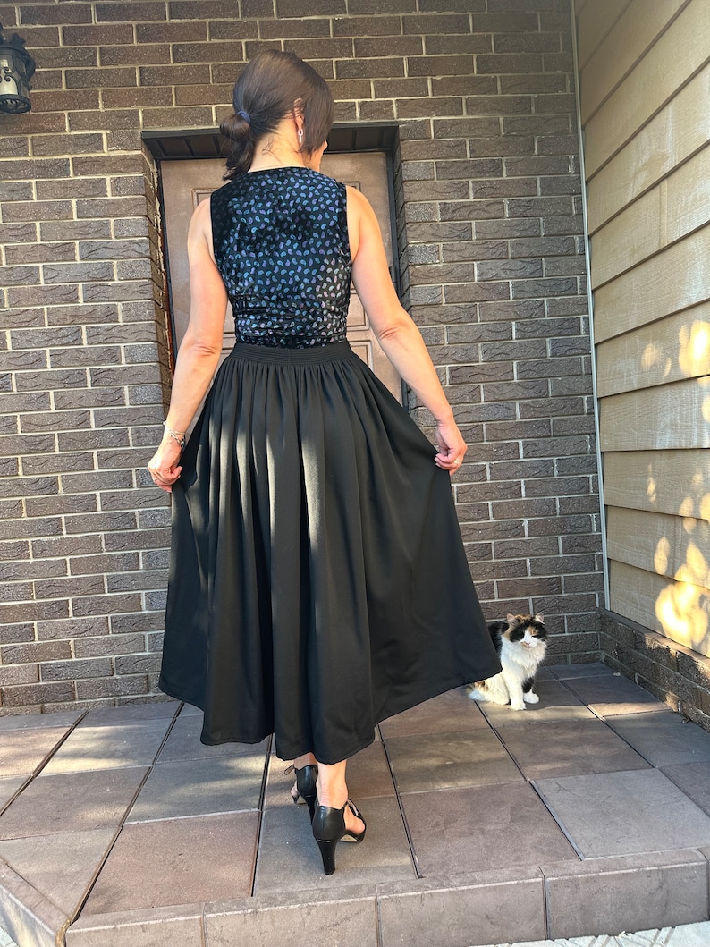 May include: A woman wearing a black skirt with a blue and black patterned top. She is standing on a tiled surface with a cat sitting in the background.