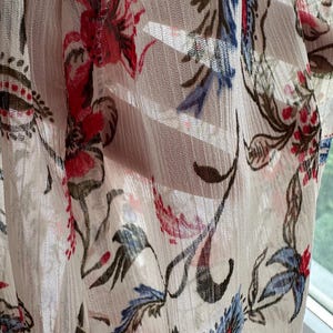 May include: A sheer white fabric with a floral pattern in shades of red, blue, and green. The fabric is draped and has a soft, flowing texture.
