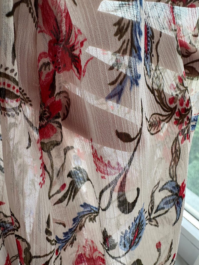 May include: A sheer white fabric with a floral pattern in shades of red, blue, and green. The fabric is draped and has a soft, flowing texture.