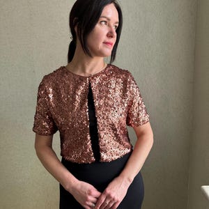 May include: A copper-coloured sequined jacket with short sleeves and an open front, worn over a black dress. The sequins give a shimmering appearance. The outfit is suitable for formal occasions or adding a touch of glamour to an ensemble.