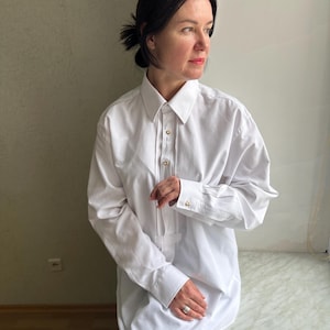 Puede incluir: Una camisa blanca de botones de gran tamaño con mangas largas y cuello. La camisa está hecha de una tela lisa y resistente a las arrugas.