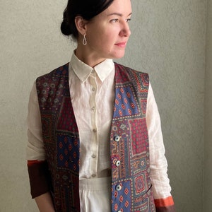 Può includere: Una donna indossa una camicia bianca abbottonata con un gilet a fantasia. Il gilet ha un design a patchwork con colori rosso, blu e oro. La donna indossa pantaloni bianchi.