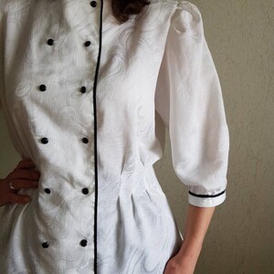 May include: White button-up blouse with a black trim and a double-breasted closure. The blouse has a slightly puffed sleeve and a fitted waist.