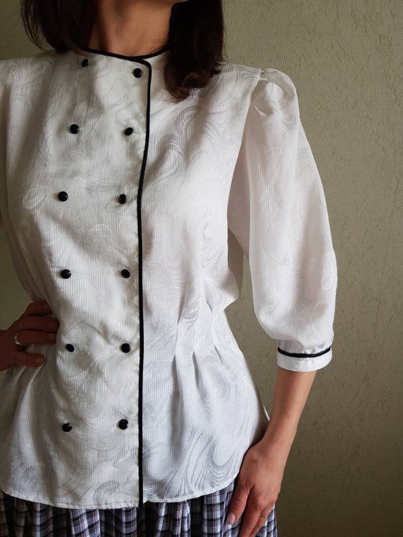 May include: White button-up blouse with a black trim and a double-breasted closure. The blouse has a slightly puffed sleeve and a fitted waist.