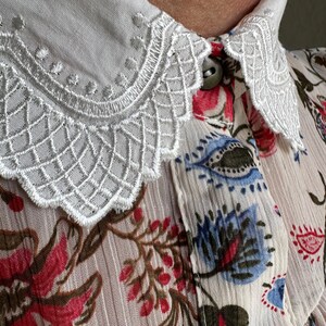 May include: A white blouse with a delicate lace collar and a floral print featuring red, blue, and green flowers on a cream background.