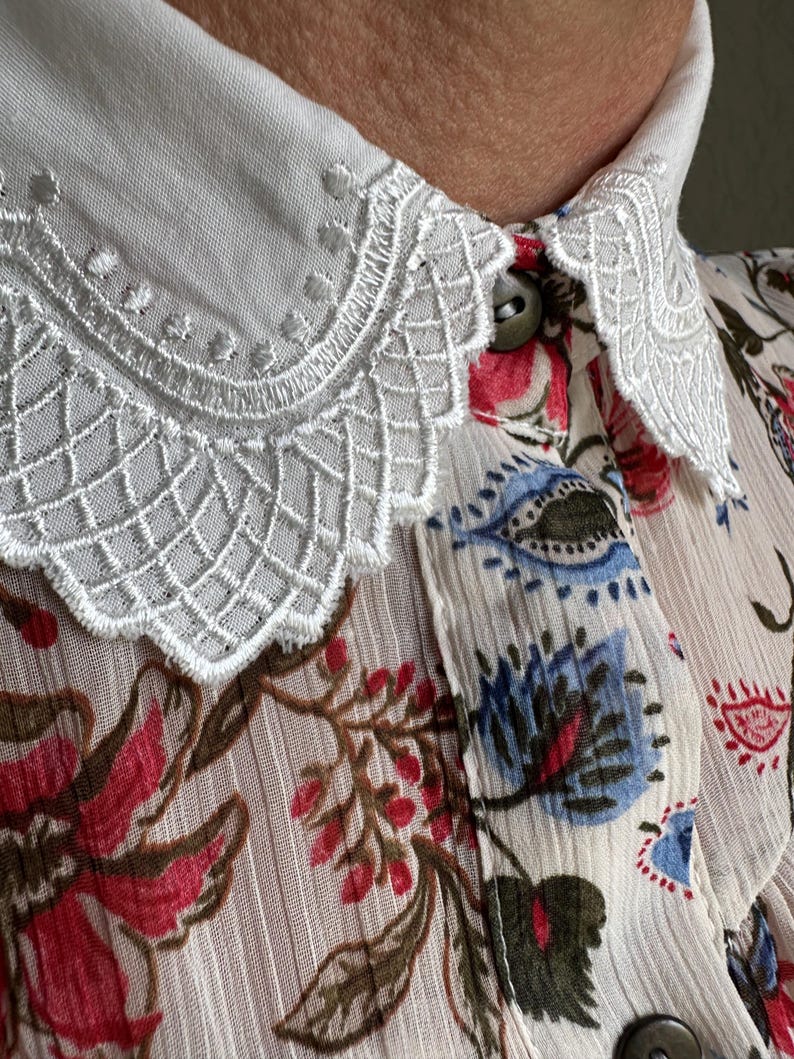May include: A white blouse with a delicate lace collar and a floral print featuring red, blue, and green flowers on a cream background.