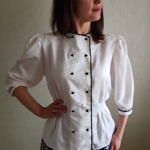 May include: White blouse with a black trim and buttons. The blouse has a fitted waist and a slightly puffed sleeve.