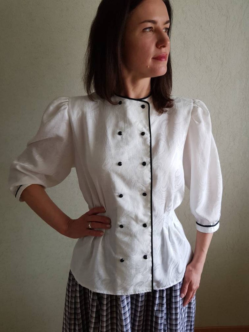 May include: White blouse with a black trim and buttons. The blouse has a fitted waist and a slightly puffed sleeve.
