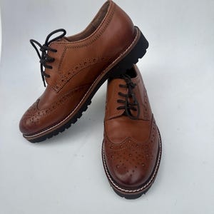 May include: Two brown leather Oxford shoes with black laces. The shoes have a classic wingtip design with decorative perforations and a thick black sole. The shoes are displayed on a white background.