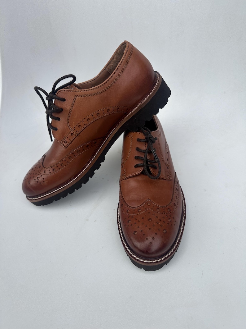 May include: Two brown leather Oxford shoes with black laces. The shoes have a classic wingtip design with decorative perforations and a thick black sole. The shoes are displayed on a white background.