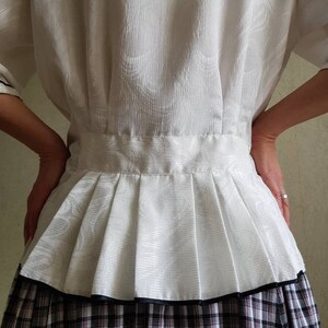 May include: A white blouse with a pleated skirt. The blouse has a textured pattern and a black trim around the bottom of the skirt. The skirt is a plaid pattern with black, white, and red.