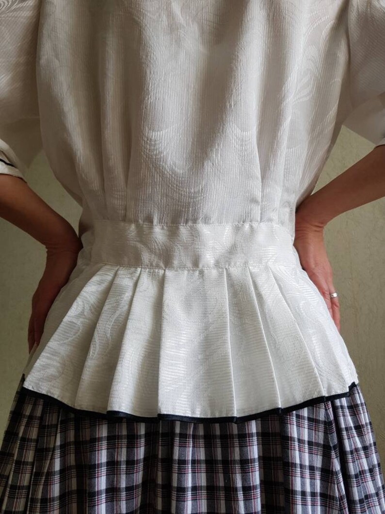 May include: A white blouse with a pleated skirt. The blouse has a textured pattern and a black trim around the bottom of the skirt. The skirt is a plaid pattern with black, white, and red.