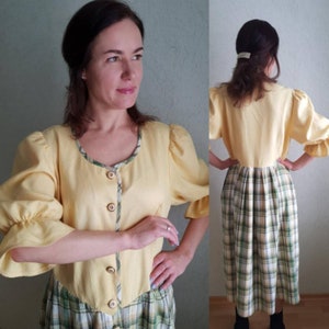 Puede incluir: Un vestido de lino amarillo con falda a cuadros. El vestido tiene un frente abotonado y mangas abullonadas.