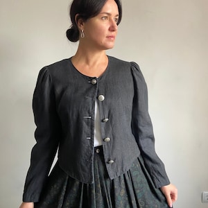 Puede incluir: Una chaqueta de lino negra con cuello redondo y cierre de botones. La chaqueta tiene una longitud recortada y mangas abullonadas. La modelo lleva una camisa blanca y una falda negra y verde.