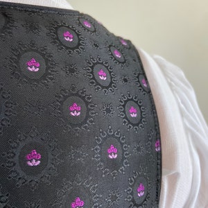 Puede incluir: Un chaleco negro con un patr&oacute;n circular repetitivo que incluye peque&ntilde;os motivos florales en magenta y morado claro. La prenda tiene un borde estructurado y se lleva sobre una camisa blanca con textura de tela visible y cuello sencillo.