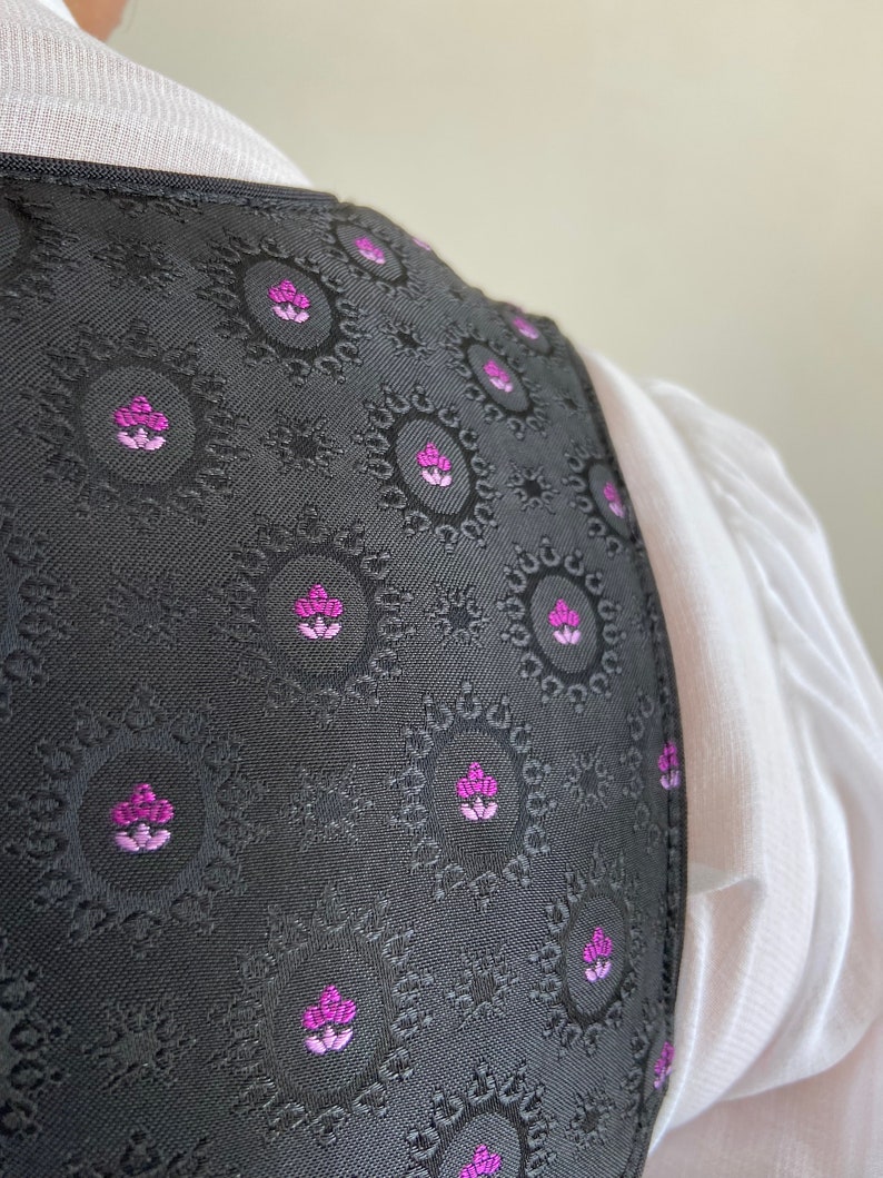 Puede incluir: Un chaleco negro con un patr&oacute;n circular repetitivo que incluye peque&ntilde;os motivos florales en magenta y morado claro. La prenda tiene un borde estructurado y se lleva sobre una camisa blanca con textura de tela visible y cuello sencillo.