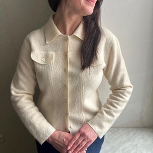 May include: Cream-colored button-down cardigan with two chest pockets. The long-sleeved sweater has a collared neckline and is made of a textured fabric. The woman is wearing a dark blue skirt or pants.