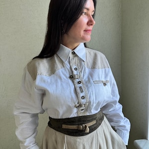 Puede incluir: Una camisa blanca de manga larga con paneles beige en los hombros y cierre de botones. La camisa se combina con un cinturón ancho marrón oscuro y una falda beige. El conjunto tiene un estilo vintage o western.