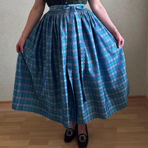 Könnte beinhalten: Ein ärmelloses, dunkelgrünes Oberteil mit einer Reihe von Knöpfen, kombiniert mit einem blau gemusterten Rock. Der Rock hat ein geometrisches Design in Blau- und Silbertönen. Schwarze Loafer mit dekorativen Schnallen vervollständigen das Outfit.