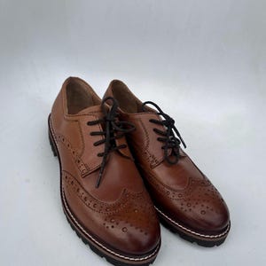 May include: A pair of brown leather brogue shoes with black laces. The shoes have decorative perforations and a thick, textured sole with a white and brown trim. The shoes are displayed on a white background.