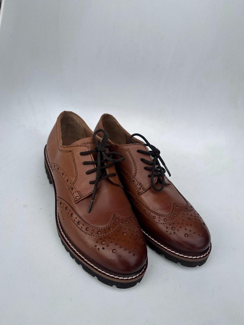 May include: A pair of brown leather brogue shoes with black laces. The shoes have decorative perforations and a thick, textured sole with a white and brown trim. The shoes are displayed on a white background.