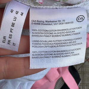 May include: A white garment care label with black text attached to a light gray fabric featuring a pink and green floral pattern. The label displays the C&A brand logo, address, and material composition, stating 100% cotton for both shell and lining.