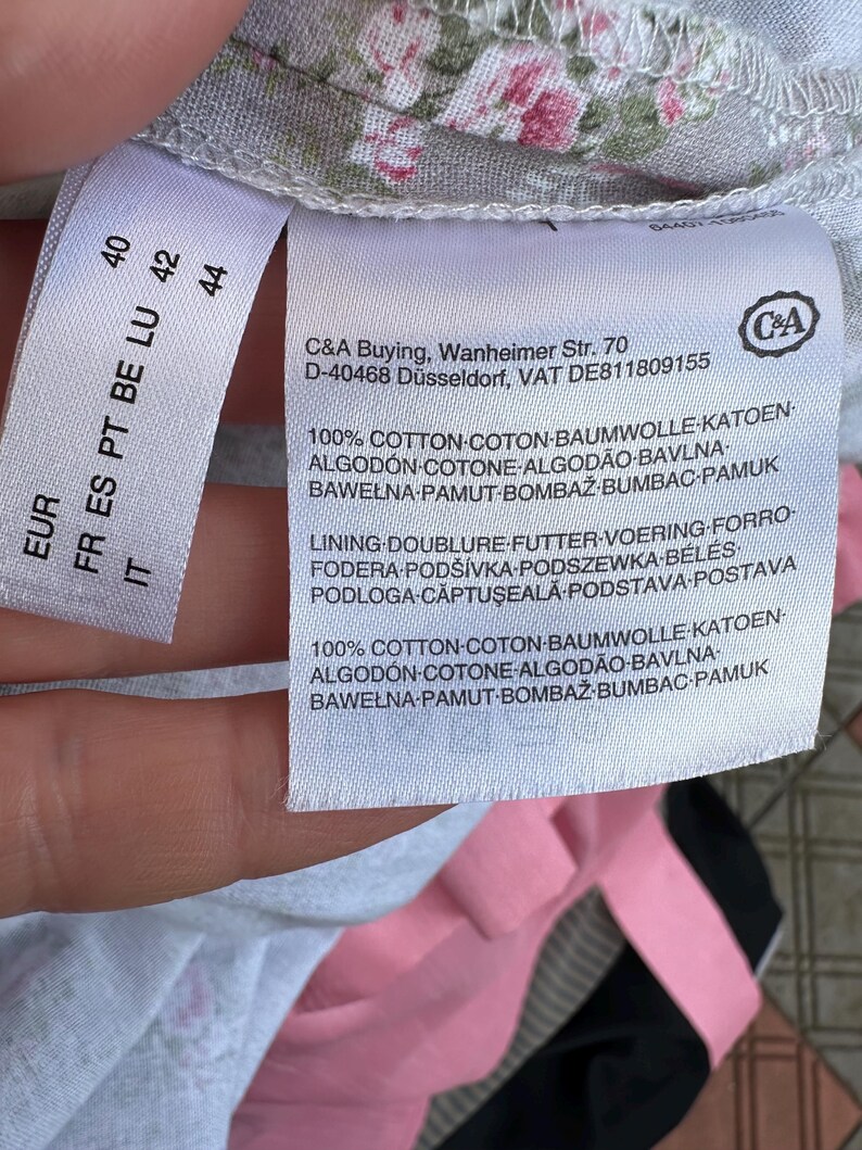 May include: A white garment care label with black text attached to a light gray fabric featuring a pink and green floral pattern. The label displays the C&A brand logo, address, and material composition, stating 100% cotton for both shell and lining.