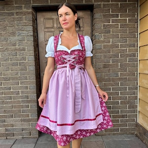 May include: A dirndl dress featuring a dark magenta bodice with a white snowflake pattern, a white blouse with short puffed sleeves, and a light lavender apron with a subtle white dot pattern and a dark magenta ribbon trim at the hem.