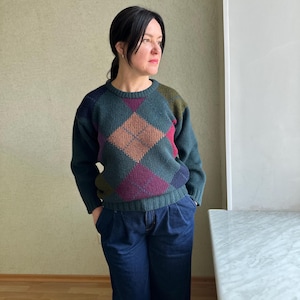 May include: A dark green argyle sweater with a crew neck and long sleeves. The argyle pattern features diamonds in shades of brown, purple, and green. The sweater is paired with dark blue denim pants.